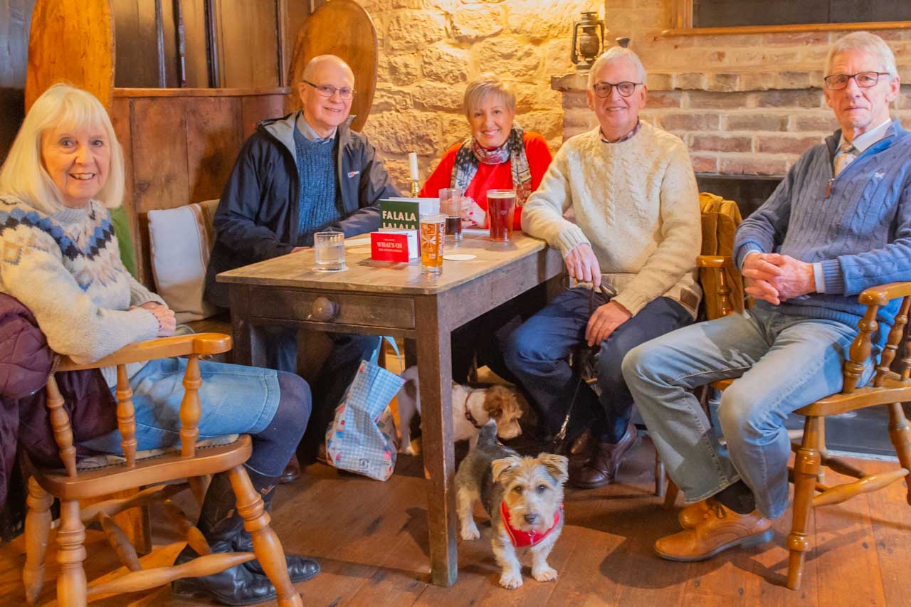 Sharing information during a meeting in the Fox & Hounds. Attendees: Heather Langford, Nigel Collins, Debbie Sutton, Debbie's partner, Roger, and Buddy Langford